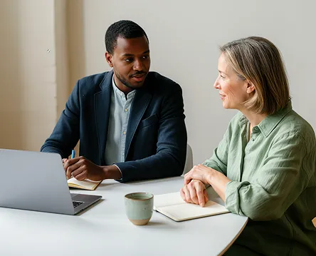 Man and woman engaged in discussion at a white table with a laptop, notebook, and coffee cup.