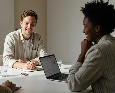 Smiling man with notebook talks with a woman during a meeting at a white table with a laptop.