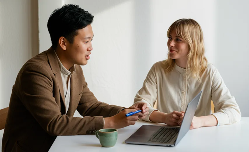 Man and woman sitting at a table engaged in conversation with a laptop and coffee cup in front of them.